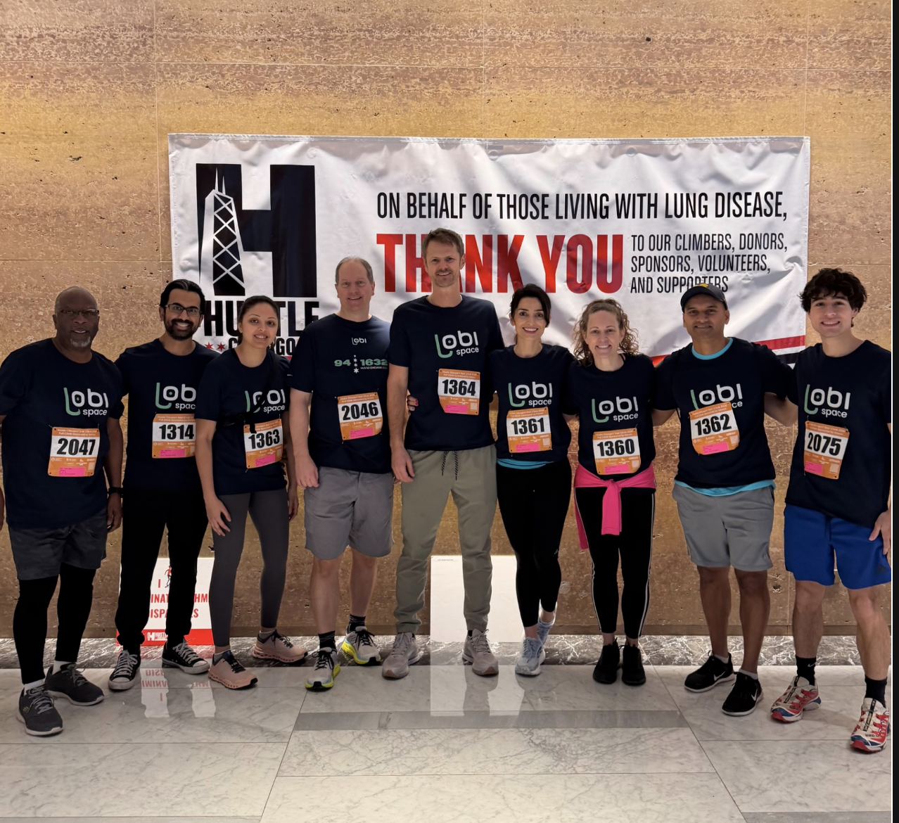 Nine members of Team Lobi Space in navy Lobi Space shirts with race bibs, standing in the John Hancock Center lobby in front of a Hustle Chicago thank you banner