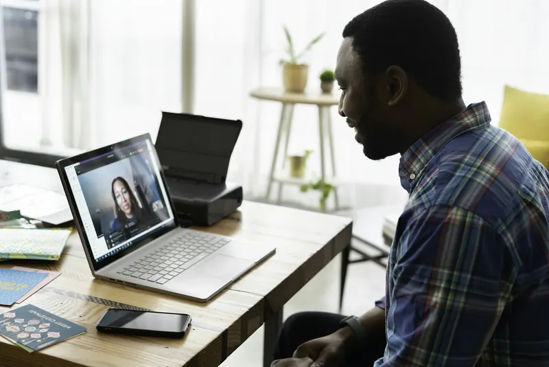 Man in a plaid shirt smiling during a video call with a colleague on his laptop at a sunny desk