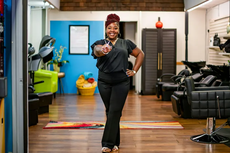 Smiling salon owner holding styling shears and pointing toward the camera in her bright hair salon